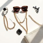 Flat lay of trending 2025 fashion accessories including a smart ring, oversized sunglasses, chunky chain necklaces, recycled handbag, and statement hair clips.