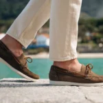 Stylish person wearing beige boat shoes and white pants walking on a boardwalk at sunset.