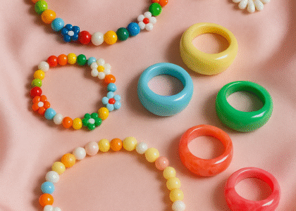 Close-up of colorful resin rings and beaded necklaces laid out on a pastel pink satin background, styled for a 2025 fashion flatlay.